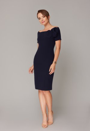 Kobieta stojąca w dopasowanej granatowej sukience z odsłoniętymi ramionami i bufkami na rękawach oraz w cielistych szpilkach na kostkę, na tle jednolitego szarego tła.