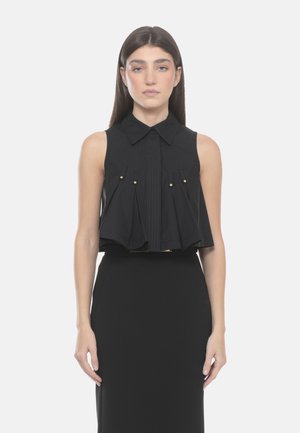 Donna con capelli castani lunghi indossa una camicetta nera senza maniche con pieghe e bottoni dorati, abbinata a una gonna nera a vita alta.