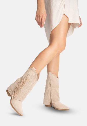 Jambes croisées portant des bottes mi-mollet beiges perforées avec des fermetures éclair latérales et des détails à pompons, associées à une robe fendue de couleur claire.