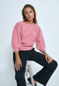 Rosa gestrickter Pullover aus strukturiertem, flauscheligem Stoff; runder Halsausschnitt; Puffärmel; kombiniert mit schwarzen weit geschnittenen Hosen.