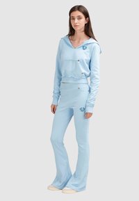 Ensemble deux pièces bleu clair comprenant un sweat à capuche court avec une poche avant et des accents en strass, associé à un pantalon évasé. Texture du tissu lisse.