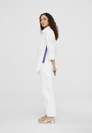 Camisa blanca oversize con mangas de tres cuartos, presenta rayas moradas y rojas en el lateral, combinada con pantalones rectos blancos y sandalias metálicas doradas.