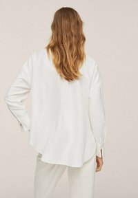Weiße Bluse mit langen Ärmeln, lockerem Schnitt, einer einfachen Naht an der Rückseite sowie seitlichen Schlitzen an den Manschetten. Glatte Textur und fließendes Design.
