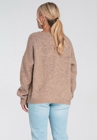 Kobieta z blond kucykiem, nosząca złote kolczyki w kształcie kół, luźny beżowy sweter dzianinowy i jasnoniebieskie dżinsy, stojąca tyłem do aparatu.