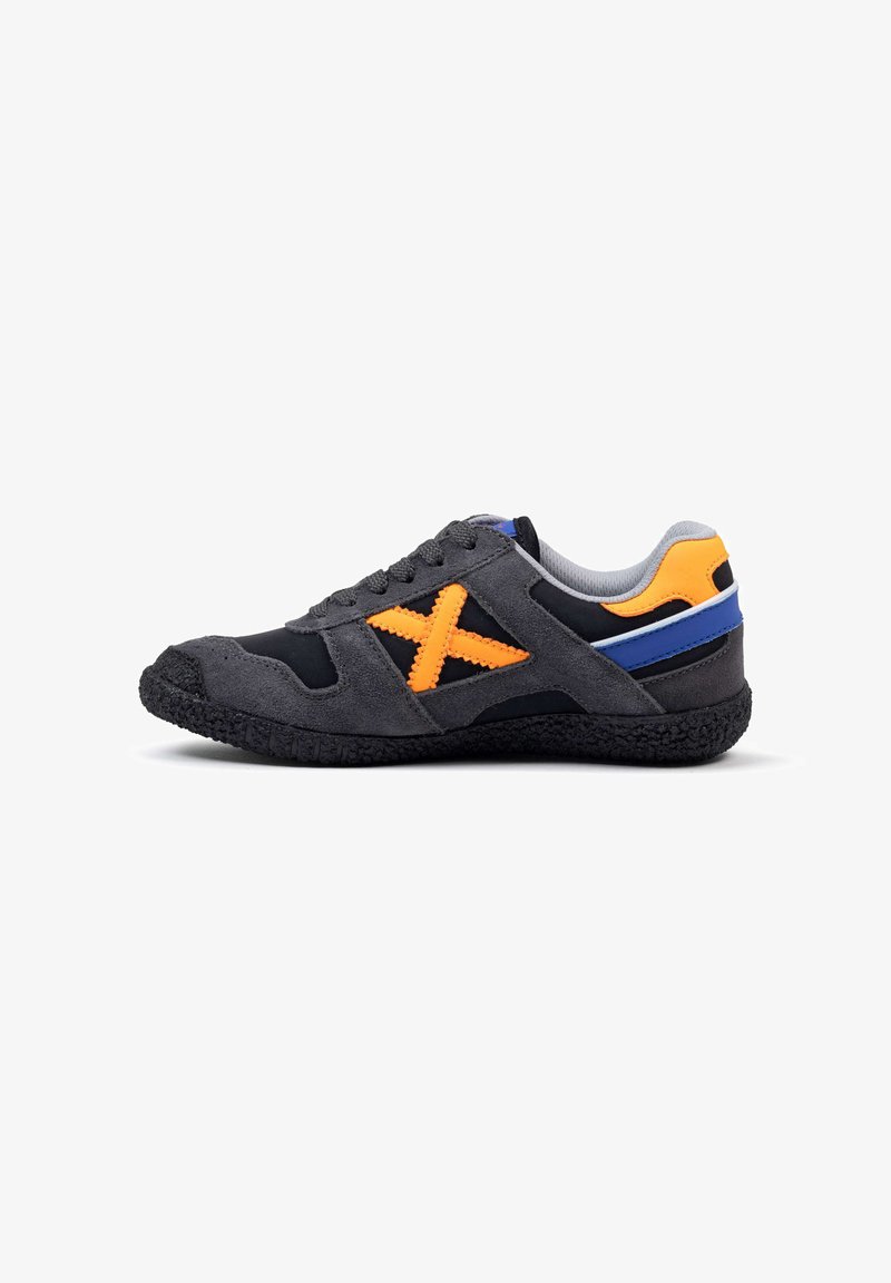Zapatilla deportiva con parte superior de ante negro, acentos en un color naranja brillante en forma de 'X', y suela de goma texturizada. Presenta un forro gris y una franja azul.