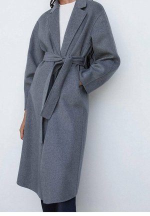 Femme portant un long manteau en laine gris ceinturé sur un haut blanc, les mains partiellement dans les poches, debout devant un fond clair uni.