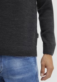 Pull en mélange de laine gris foncé avec un ourlet et des poignets côtelés, finition texturée et une petite étiquette de marque sur le côté. Porté avec un jean bleu.
