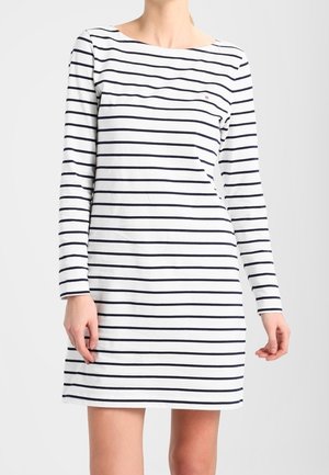 Femme portant une robe blanche à manches longues avec des rayures noires horizontales espacées de manière régulière, debout devant un fond clair uni.