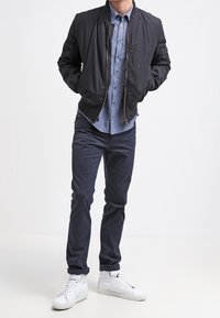 Black bomber jacket with a zip front, paired with a light blue button-up shirt, navy pants, and white high-top sneakers.