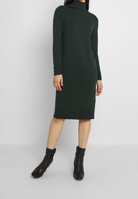 Långärmad, mörkgrön stickad klänning med hög krage, kroppsnära silhuett och ribbade ärmar, tillsammans med svarta ankelskor.