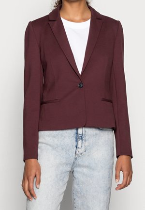 Person iført en tætsiddende mørk bordeaux blazer over en hvid skjorte og lyseblå jeans, stående med armene afslappet ned langs siderne.