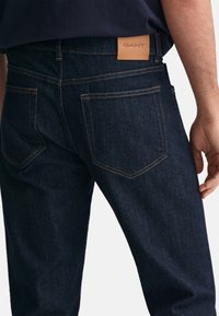 Jeans in denim blu scuro con una piccola toppa in pelle marrone chiaro sulla vita posteriore, dotati di due tasche posteriori e dettagli di cucitura a contrasto.