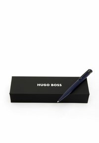 Stylo à bille bleu sur une boîte noire avec "HUGO BOSS" imprimé en blanc. Le stylo a une finition lisse avec un clip et une pointe arrondie.