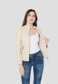 Licht beige nepleren jas met ritssluiting, doorgestikte schouders en twee zijzakken. Gecombineerd met blauwe spijkerbroek met bloemmotieven.