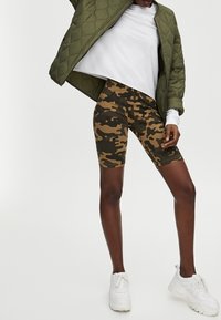 Bermudashort in camouflagepatroon met tinten bruin en groen, gecombineerd met een wit shirt met lange mouwen en een donkergroene doorgestikte jas.