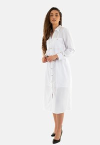 Robe blanche à boutons avec col, deux poches poitrine, accents en or, et coupe droite. Portée avec des escarpins noirs.