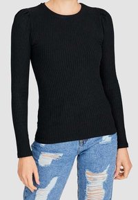 Kvinna som bär en åtsittande svart ribbad långärmad top tillsammans med högmidjade blå slitna jeans mot en enkel bakgrund.