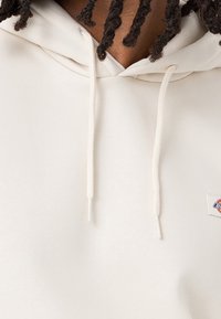 Krémová mikina s měkkou texturou, vybavená šňůrkami a malou logo nášivkou. Design zahrnuje volný střih.