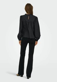 Blouse noire à manches longues avec des manches bouffantes, un col volanté et un détail en forme de trou de serrure au dos, associée à un jean noir évasé et des chaussures à talons.