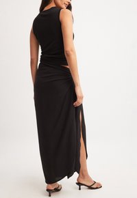 Top senza maniche nero abbinato a una lunga gonna nera con spacco laterale, texture liscia e design semplice. Sandali con tacco eleganti completano l'outfit.