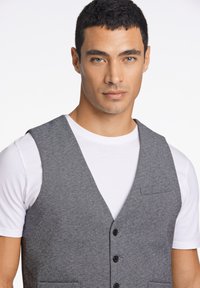 Grå tyggvest med en strukturerad yta, med V-hals, fem knappar och en bröstficka, bärs över en vit t-shirt.