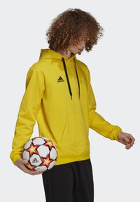 Gul pullover hoodie med sorte snore og logo, der har en frontlomme. Personen holder en fodbold med røde og gule accenter.