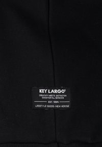 Fekete szövet, amelyen a "KEY LARGO" szöveget tartalmazó címke látható, amely a márka jellemzőit is ismerteti. A címkén ábrázolva van egy bejegyzett védjegy szimbólum.