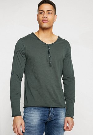 Camisa henley de manga larga en verde oliva con cuello redondeado, tapeta de seis botones y tejido suave al tacto. Combinada con vaqueros azules.