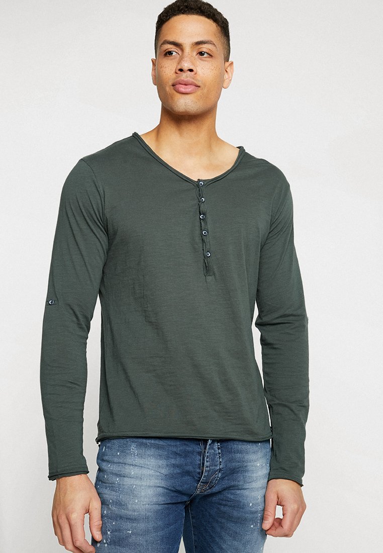 Langarm-Henley-Shirt in Olivgrün mit rundem Halsausschnitt, sechs Knöpfen und weichem, strukturiertem Stoff. Getragen mit blauen Jeans.
