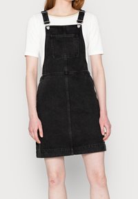 Black denim overall dress with adjustable straps, front pocket, and a straight-cut design. Features a faded texture and minimal stitching.