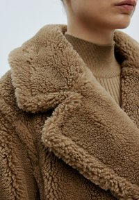 Brauner Kunstfellmantel mit einem breiten Kragen, strukturiertem Stoff und einem gemütlichen Design, kombiniert mit einem gerippten braunen Pullover darunter.