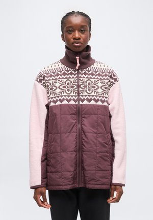 Jeune personne aux cheveux tressés portant une veste zippée à motif violet et rose avec la moitié inférieure matelassée et des manches en polaire sur un fond uni.