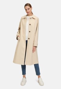 Trench coat beige con colletto, chiusura frontale a bottoni e tasche laterali. Realizzato in tessuto liscio, presenta una vestibilità dritta e maniche lunghe.
