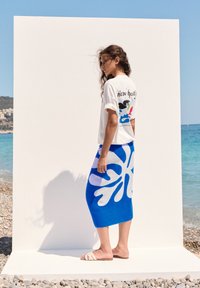 Hvid t-shirt med farverig grafik på ryggen, parret med en blå nederdel med et hvidt bladmønster. Modellen bærer beige sandaler, sat mod en strandbaggrund.