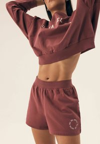 Burgundowa, skrócona bluza z krótkim rękawem oraz dopasowane szorty, wykonane z miękkiego materiału. Szorty mają elastyczny pas i okrągły napis graficzny.