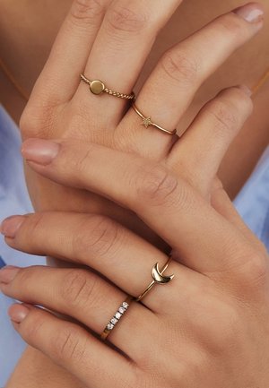 Mains avec quatre bagues en or : anneau torsadé avec disque rond, anneau fin avec étoile, anneau fin avec croissant de lune, et anneau avec cinq petits diamants.