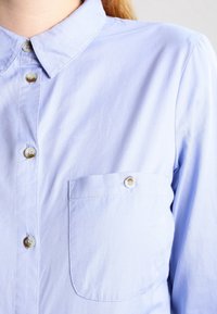 Ljusblå, krage skjorta i slätt tyg, med knappstängning fram och en enda bröstficka med knappstängning.