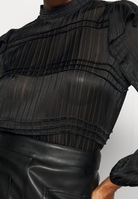 Haut noir transparent à rayures texturées, manches à volants et col montant, associé à une jupe en cuir noire taille haute.