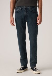 Σκούρο μπλε τζιν με ίσια εφαρμογή, σχέδιο με πέντε τσέπες, ελαφρύ ξεβάψιμο και διακριτικές λεπτομέρειες ραφής, συνδυασμένο με μαύρα αθλητικά παπούτσια.