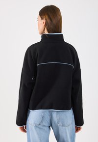 Pulover negru din polar cu guler înalt, cu accente bleu deschis pe umeri și pe tiv. Croi lejer, cu mâneci lungi și o textură netedă.