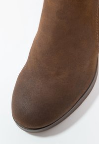 Brązowy zamszowy botek sięgający kostki z zaokrąglonym noskiem, gładką fakturą i płaską podeszwą. Projekt charakteryzuje się subtelnym przeszyciem wzdłuż boku.