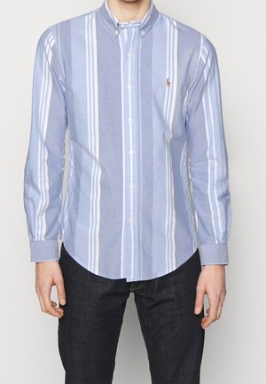 Camisa de botones de manga larga a rayas en azul claro y blanco, con collar, yugo trasero europeo, y un pequeño logo en el pecho.