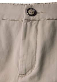 Un pantalon en coton beige présente une texture lisse, une fermeture à un bouton et des coutures le long de la zone des poches avant.
