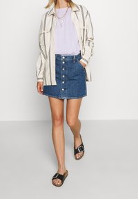 Mini-jupe en denim avec fermeture à boutons sur le devant, associée à un t-shirt violet clair et une surchemise beige à rayures ; sandales noires à glissière avec boucle.