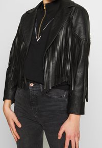 Czarna kurtka z ekologicznej skóry z frędzlami, kołnierzem z klapami i zamkiem na przodzie, z długimi frędzlami, noszona na czarnej bluzce i dżinsach.