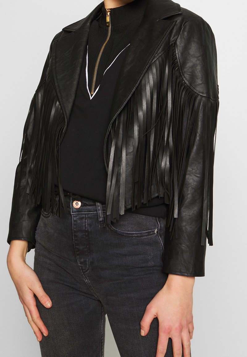 Czarna kurtka z ekologicznej skóry z frędzlami, kołnierzem z klapami i zamkiem na przodzie, z długimi frędzlami, noszona na czarnej bluzce i dżinsach.
