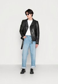 Zwart leren motorjack, wit T-shirt, lichtblauwe opgerolde spijkerbroek en zwarte schoenen. Model draagt een zonnebril. Minimalistische achtergrond.