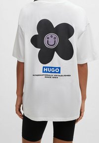 Biała, oversize'owa koszulka z dużym czarnym kwiatem z lawendowym środkiem oraz napisem "HUGO INTERNATIONALLY ESTABLISHED SINCE 1993."