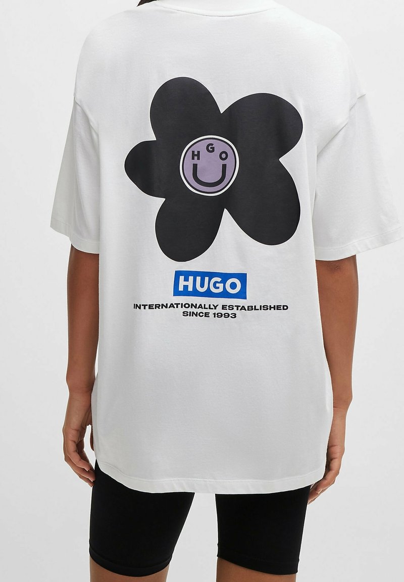 Biała, oversize'owa koszulka z dużym czarnym kwiatem z lawendowym środkiem oraz napisem "HUGO INTERNATIONALLY ESTABLISHED SINCE 1993."