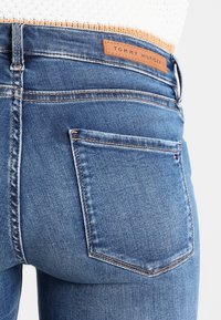 Vaqueros de mezclilla en azul lavado medio con una etiqueta de cuero de marca. Presentan un diseño clásico de cinco bolsillos y un detalle sutil de desgaste en forma de rayas.